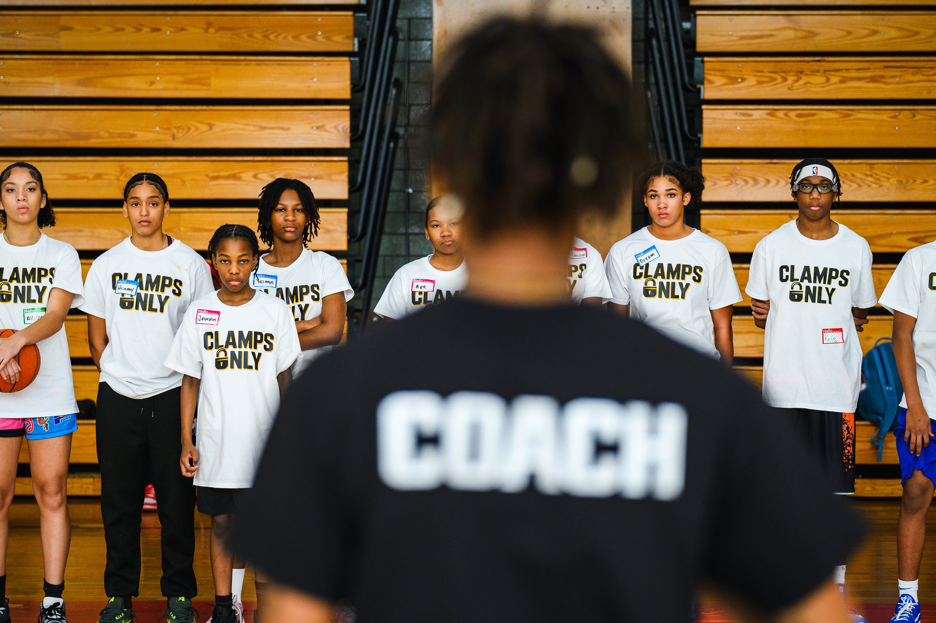A group of young basketball players wearing white t-shirts with 'CLAMPS ONLY' printed on the front, standing on a wooden gymnasium court. In the foreground, the back of a coach's head is visible wearing a black t-shirt with 'COACH' printed in large white letters.